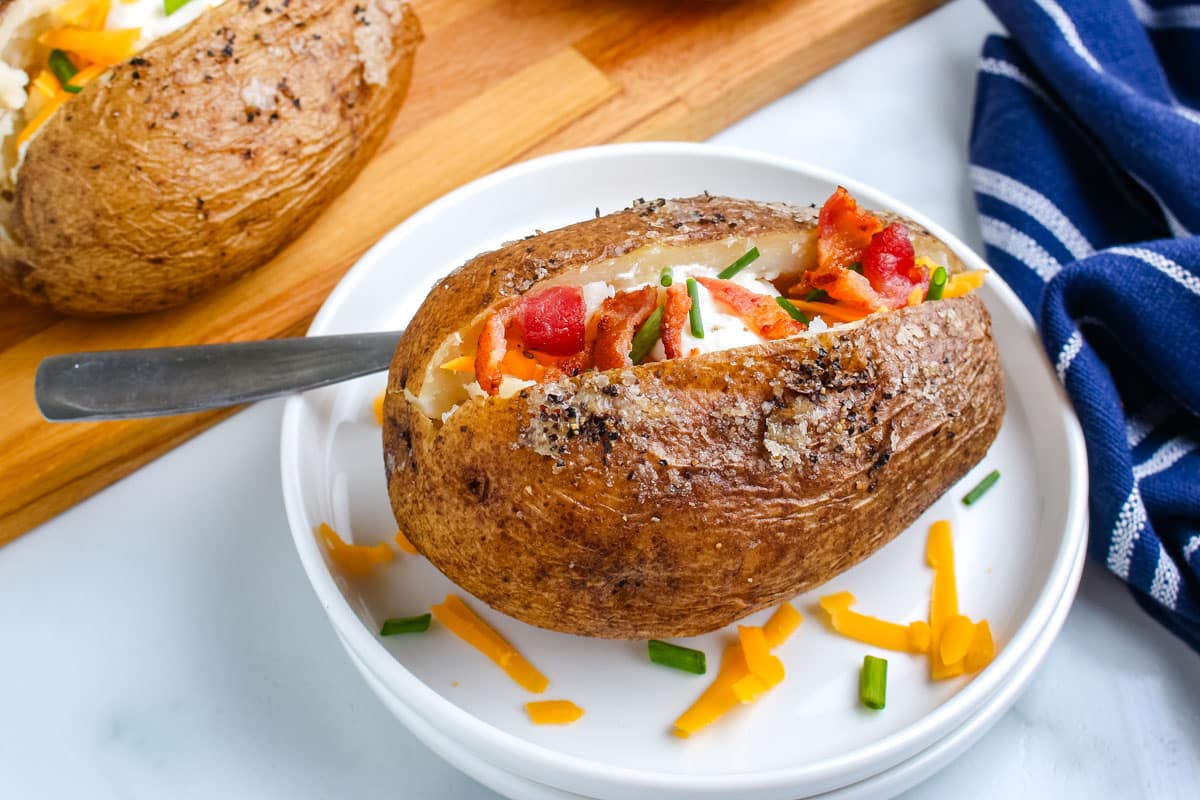 A smoked, baked potato on a white plate with cheese, butter, chives, bacon, and sour cream.
