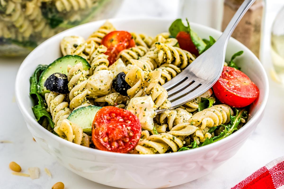 A fork diggin into the finished Pesto Pasta Salad recipe that's in a white bowl.