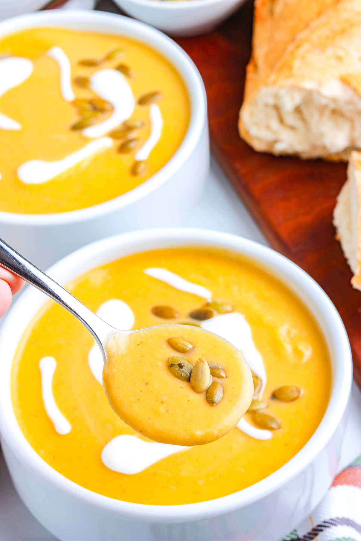 A spoonful of creamy roasted pumpkin soup topped with toasted pumpkin seeds, held over a bowl of soup drizzled with cream and served with crusty bread.