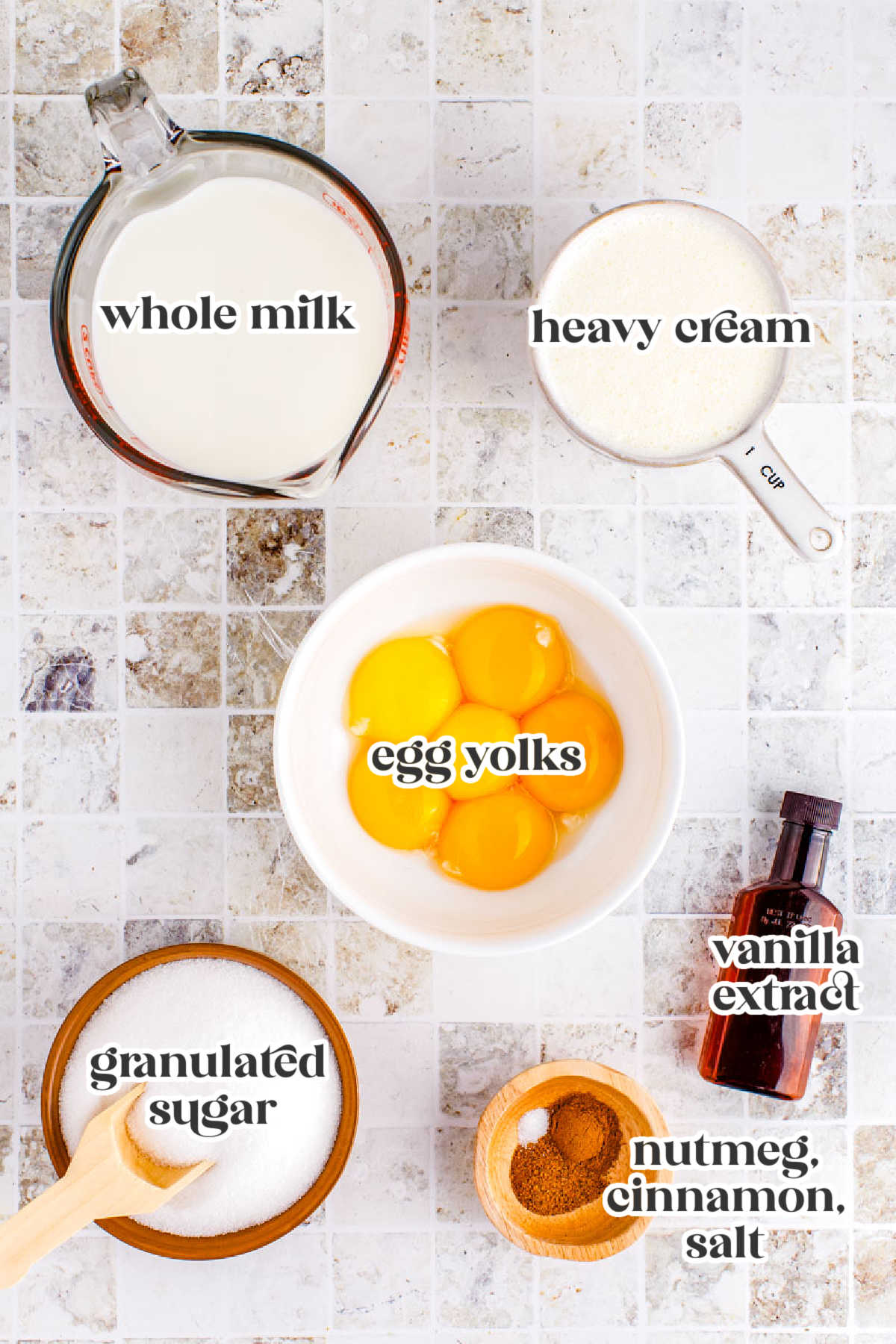 Overhead photo of homemade eggnog ingredients on a tiled counter, including whole milk, heavy cream, egg yolks, granulated sugar, vanilla extract, nutmeg, cinnamon, and salt.