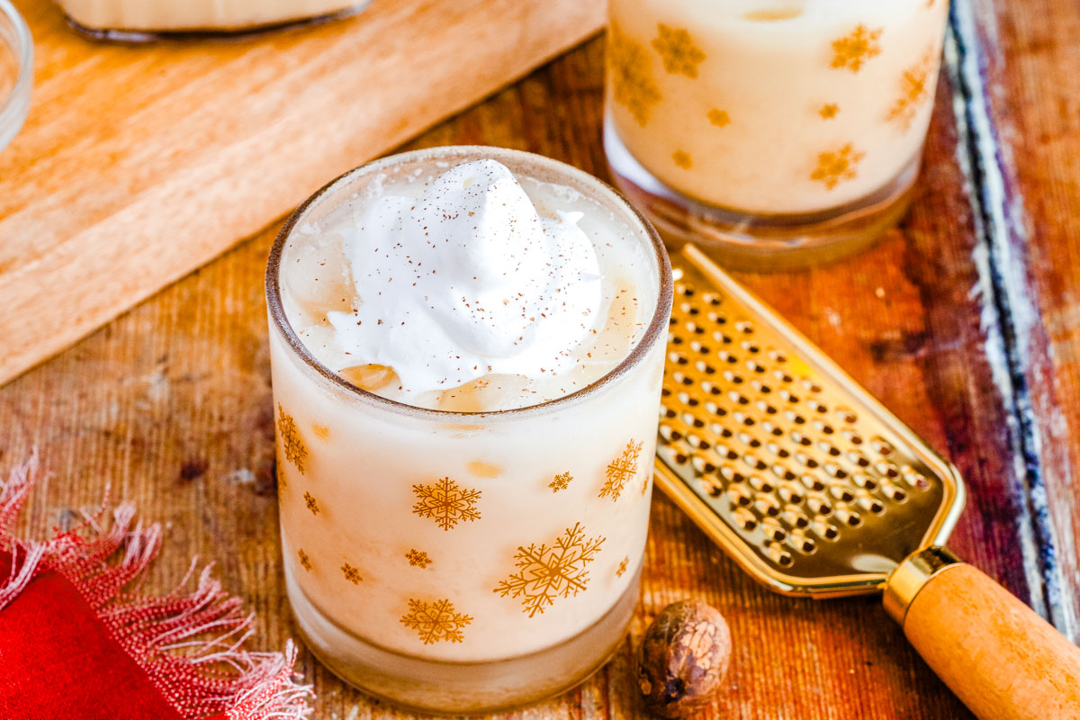 Two glasses of chilled homemade eggnog topped with whipped cream and nutmeg, with a gold grater and festive red napkin on a rustic wooden table.