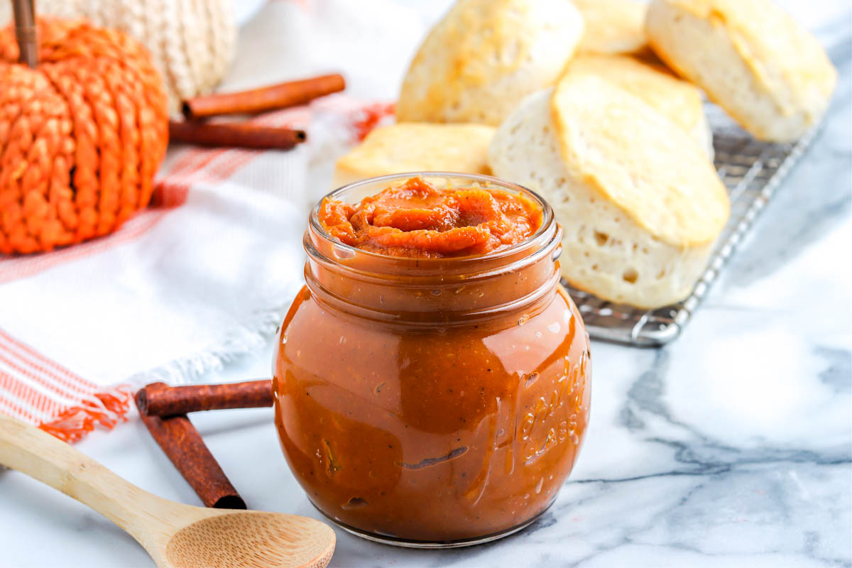 Glass jar filled with creamy spiced pumpkin butter, surrounded by biscuits and cinnamon sticks for a cozy fall spread.