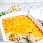 Leftover turkey casserole in a white baking dish with a cheesy golden top, partially served with a spoon showing creamy layers of turkey, vegetables, and mashed potatoes.