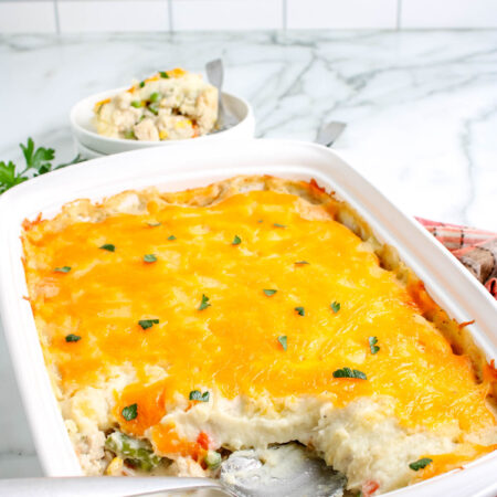 Leftover turkey casserole in a white baking dish with a cheesy golden top, partially served with a spoon showing creamy layers of turkey, vegetables, and mashed potatoes.