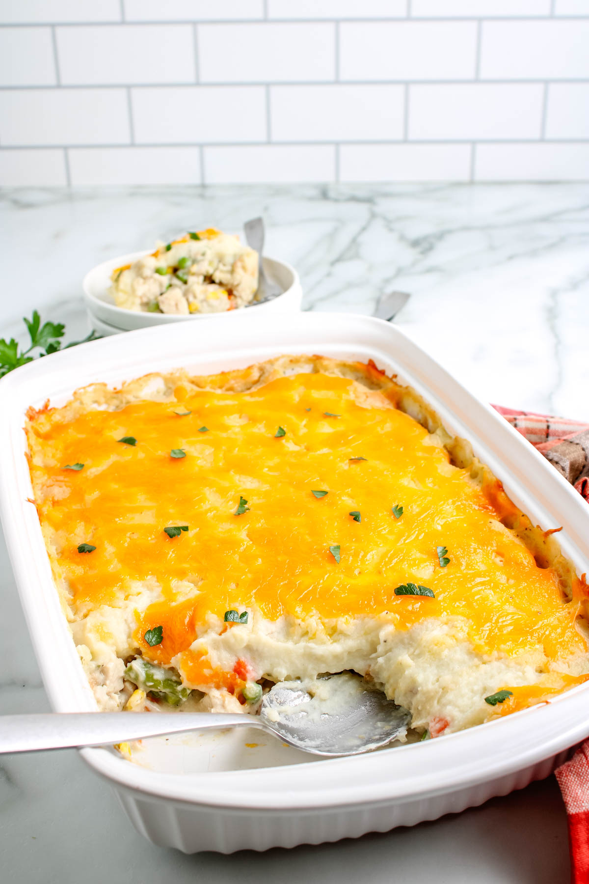 Leftover turkey casserole in a white baking dish with a cheesy golden top, partially served with a spoon showing creamy layers of turkey, vegetables, and mashed potatoes.