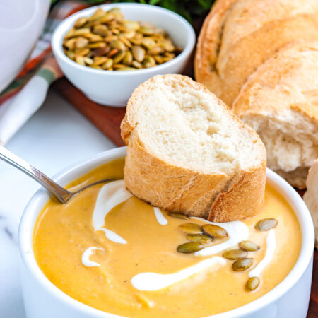 Bowl of roasted pumpkin soup garnished with cream and pumpkin seeds, with a slice of crusty bread resting on top and parsley in the background.