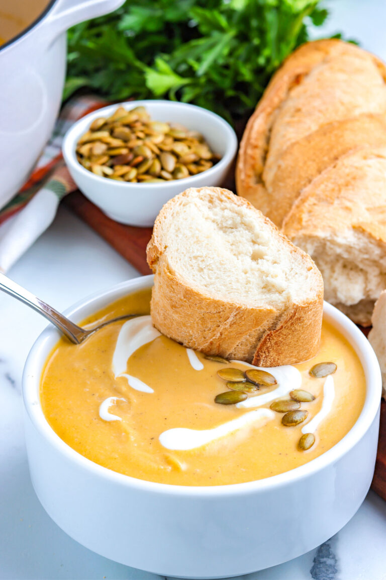 Bowl of roasted pumpkin soup garnished with cream and pumpkin seeds, with a slice of crusty bread resting on top and parsley in the background.