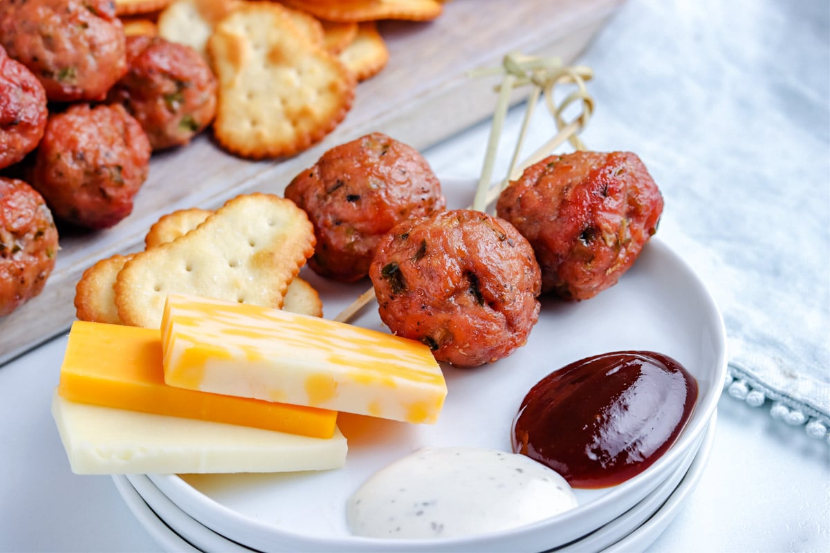 Three smoked meatballs on a plate with BBQ sauce, cheese, and crackers