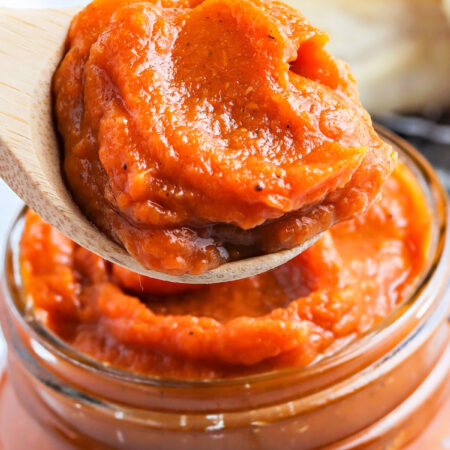 lose-up of a wooden spoon lifting thick, glossy spiced pumpkin butter from a jar.