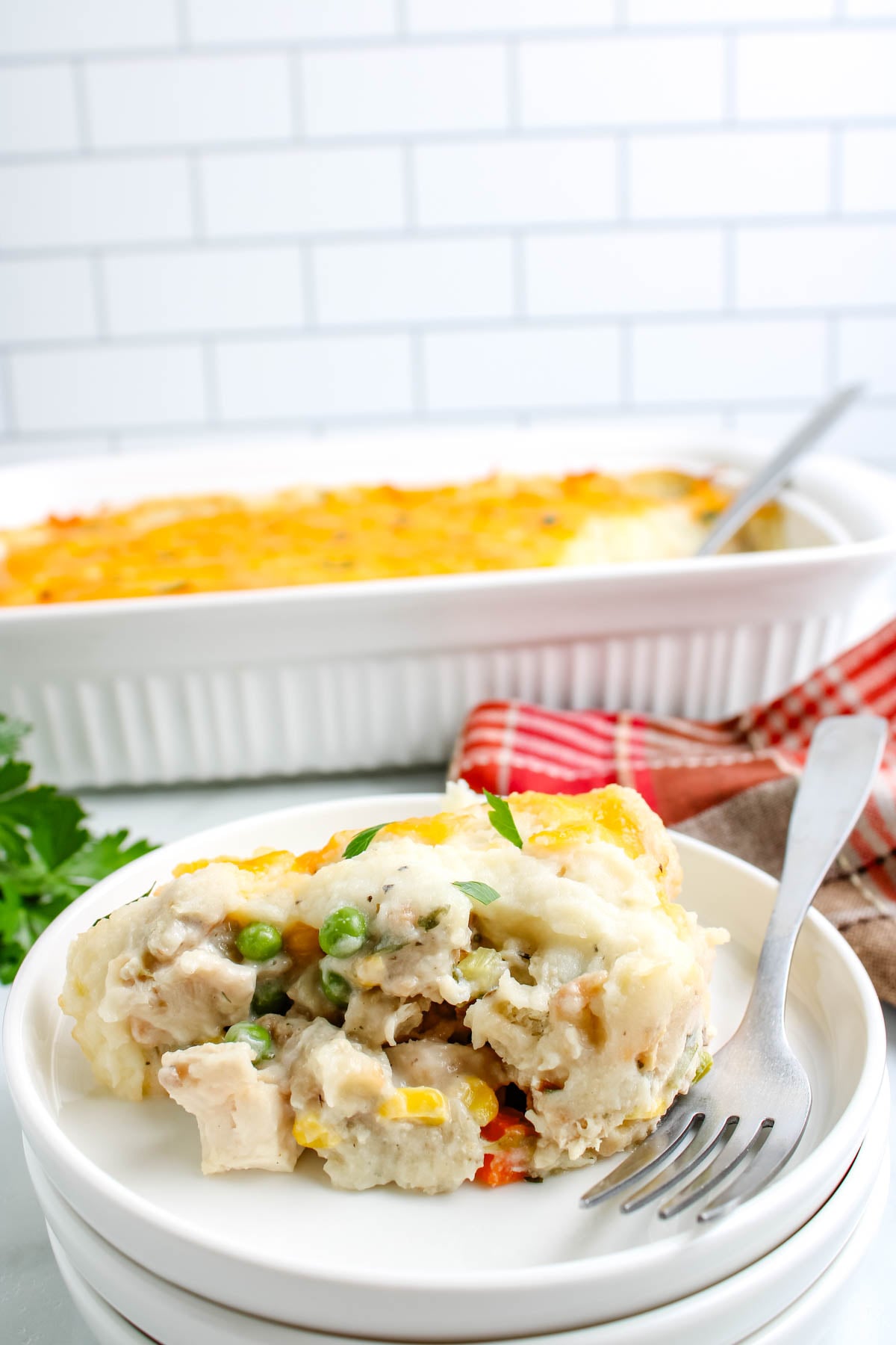 Plate with a serving of leftover turkey casserole in front of the full cheesy casserole dish, garnished with parsley and set against a kitchen tile background.