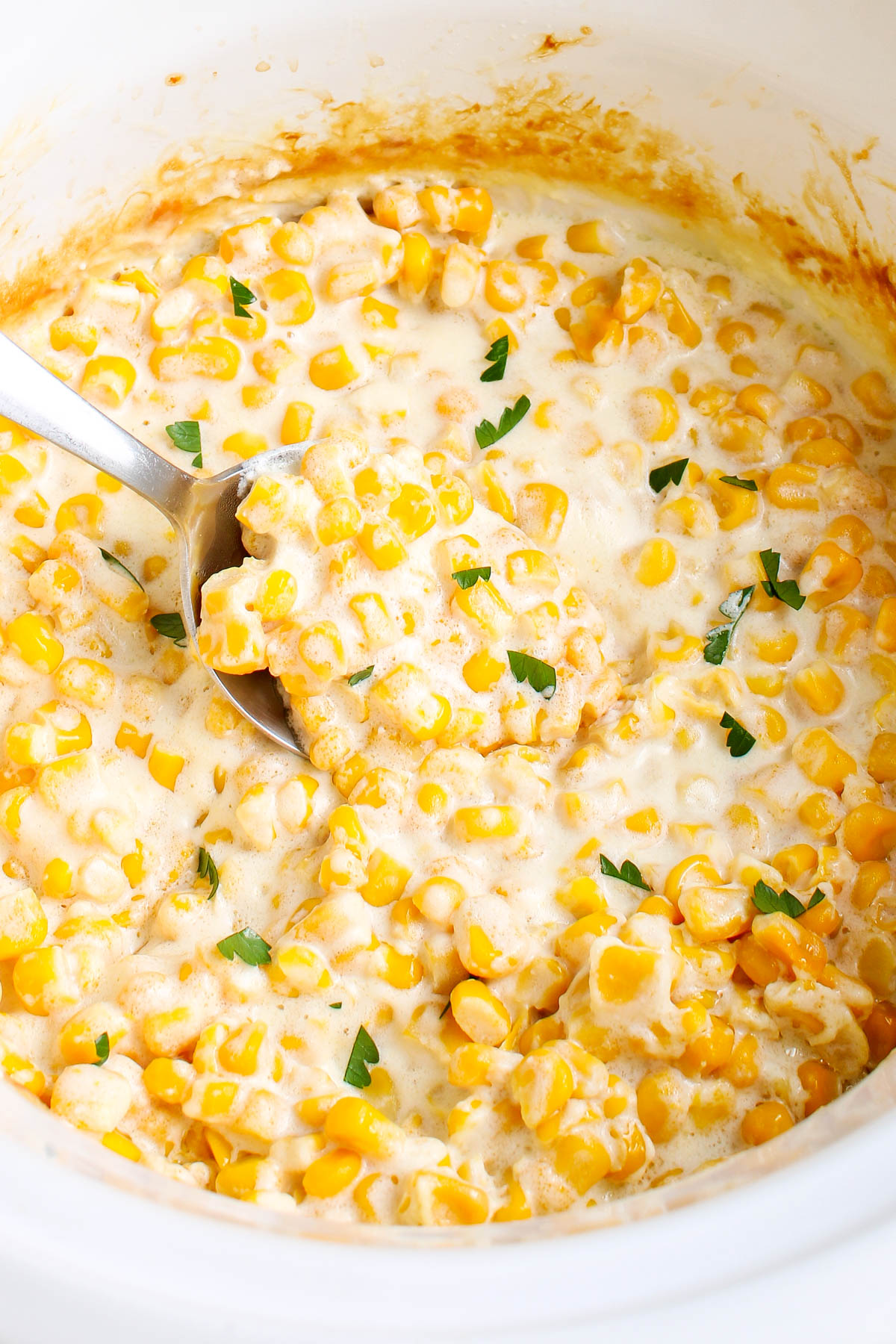 Close-up of creamy crockpot creamed corn with melted cream cheese and butter, sprinkled with fresh parsley and scooped with a serving spoon.