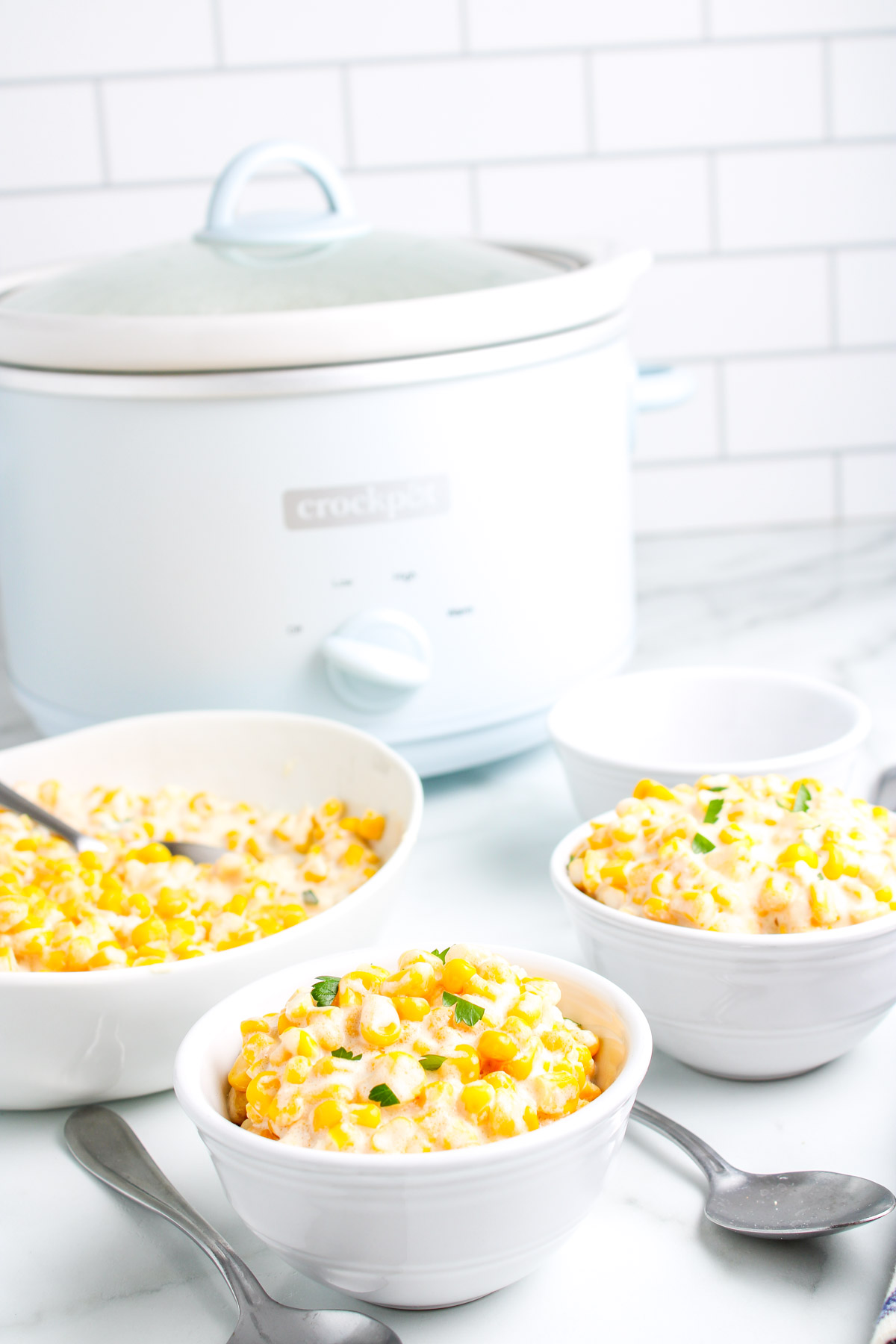 Bowls of creamed corn served beside a white crockpot on a marble counter, showing the creamy texture and golden corn kernels ready to eat.