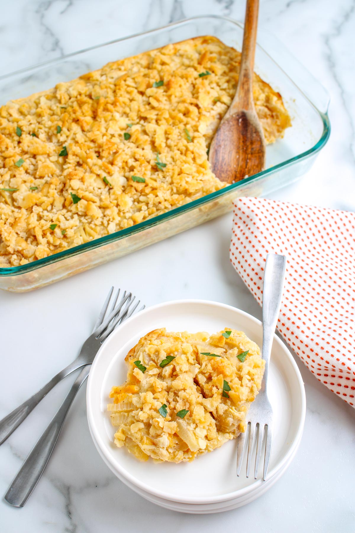 Glass baking dish of golden onion casserole with a serving scooped onto a plate beside a red and white kitchen towel.