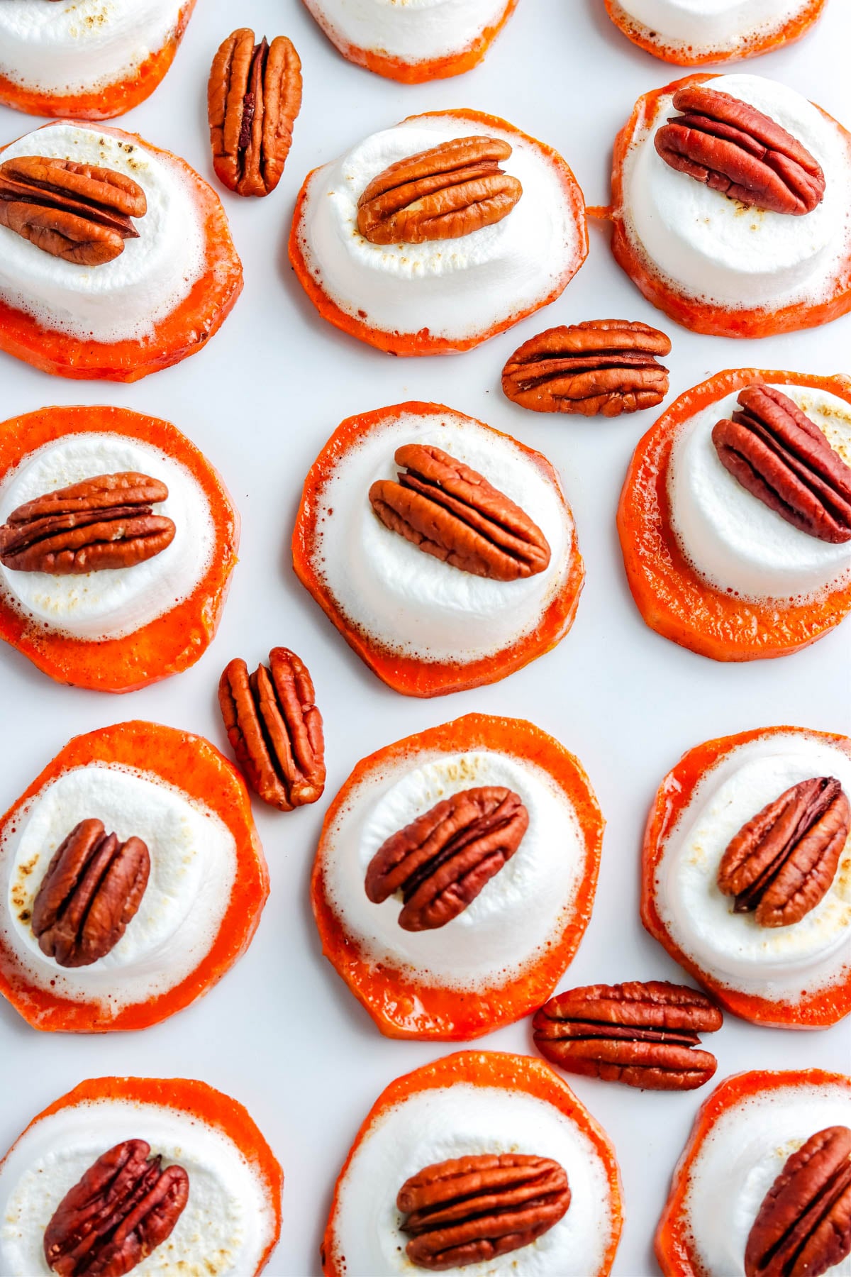 Overhead shot of sweet potato bites topped with toasted marshmallows and pecans arranged in neat rows on a white background.