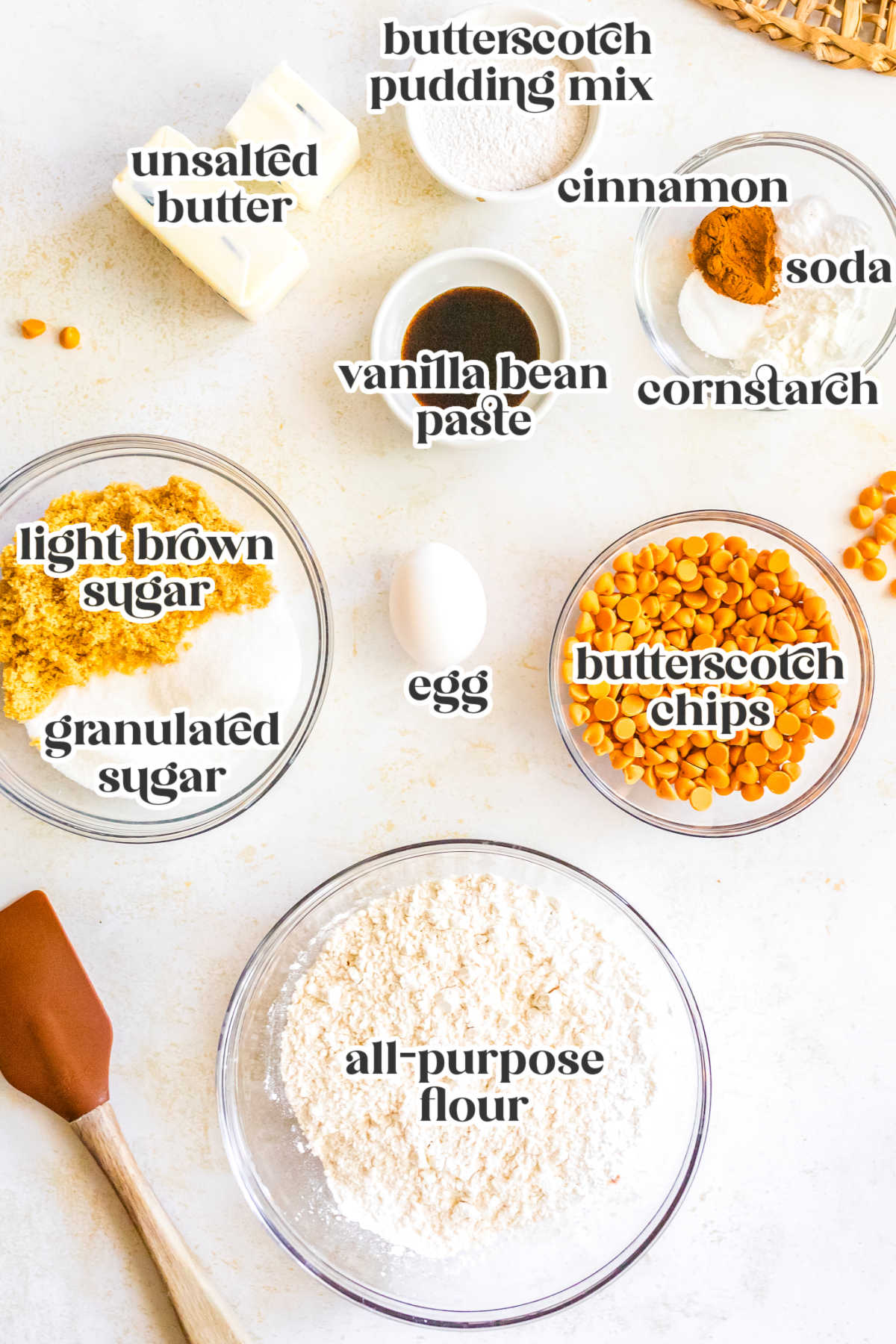 Overhead view of labeled baking ingredients for chewy butterscotch cookies including butter, pudding mix, sugars, egg, flour, cinnamon, cornstarch, baking soda, vanilla, and butterscotch chips.
