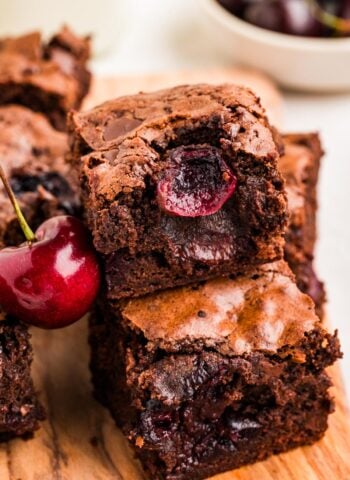 Close up view of fudgy cherry brownies stacked with juicy baked cherries inside and a fresh cherry beside them.