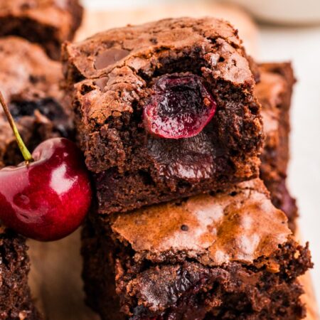 Close up view of fudgy cherry brownies stacked with juicy baked cherries inside and a fresh cherry beside them.