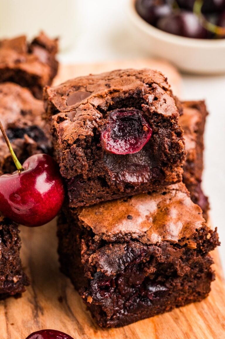 Close up view of fudgy cherry brownies stacked with juicy baked cherries inside and a fresh cherry beside them.