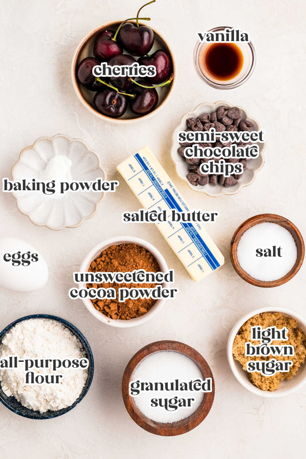 Overhead view of cherry brownie ingredients including fresh cherries, cocoa powder, flour, sugars, butter, baking powder, chocolate chips, eggs, salt, and vanilla arranged on a light surface.