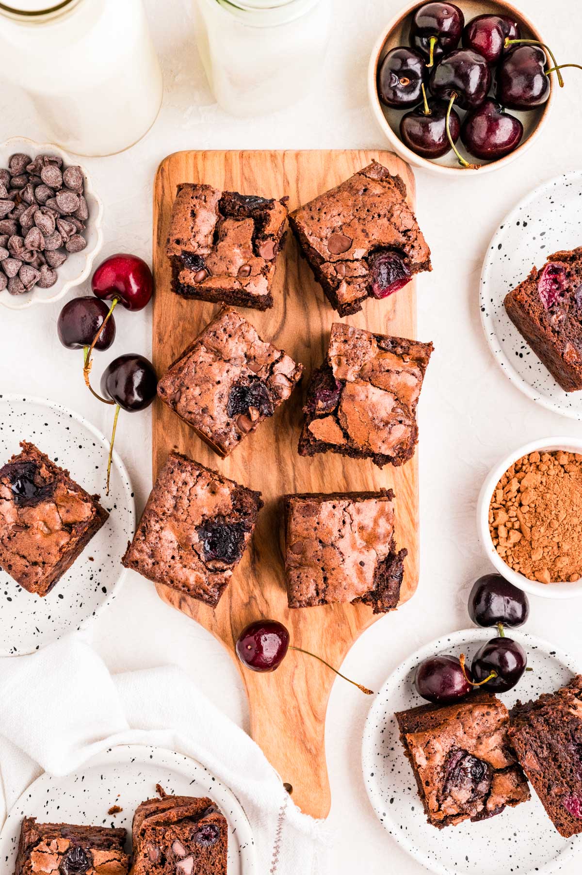 Cherry brownies sliced into squares on a wooden board surrounded by fresh cherries, chocolate chips, and cocoa powder.