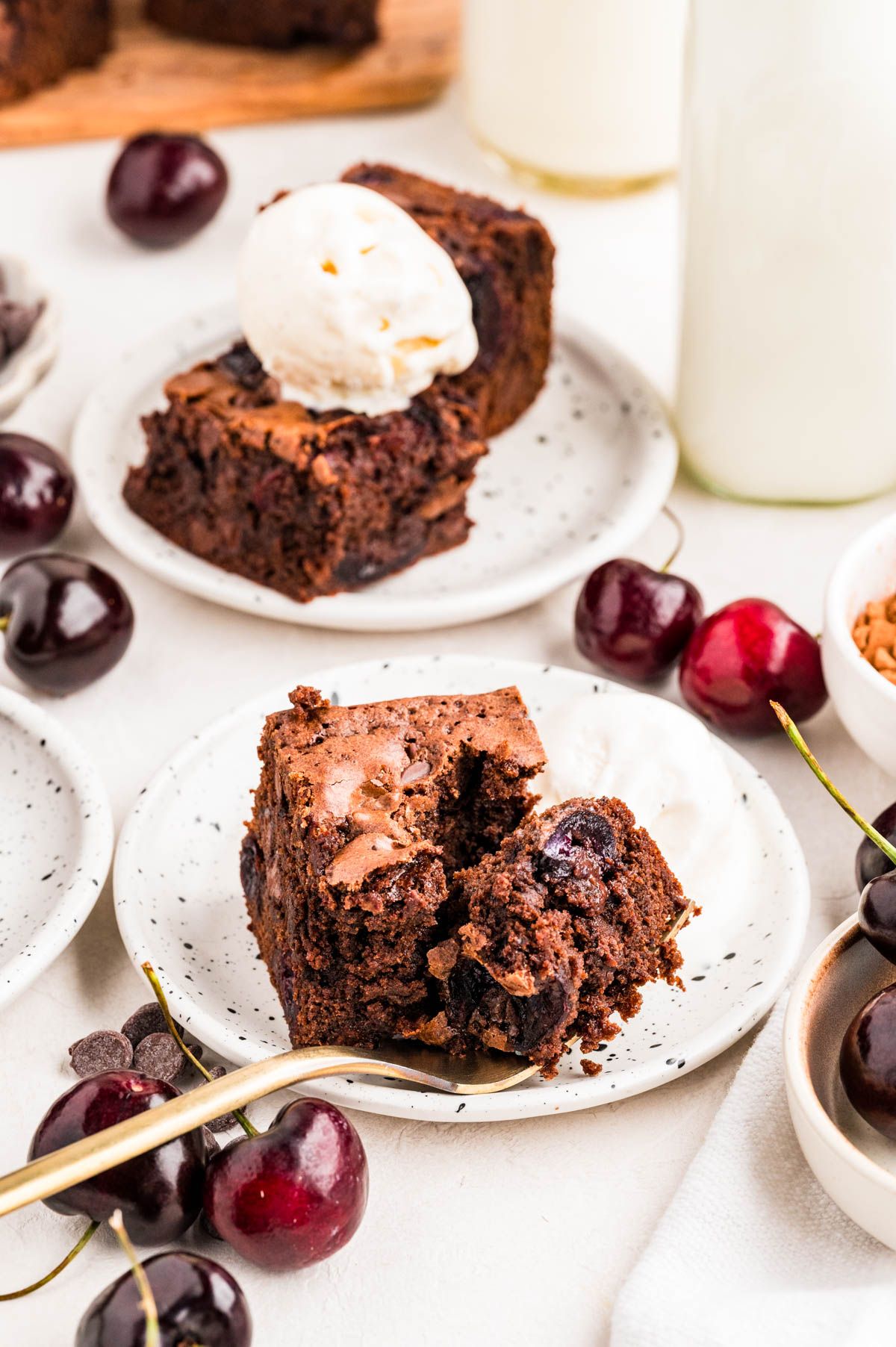 A square of cherry brownie on a speckled plate topped with a scoop of vanilla ice cream with fresh cherries around the plate.