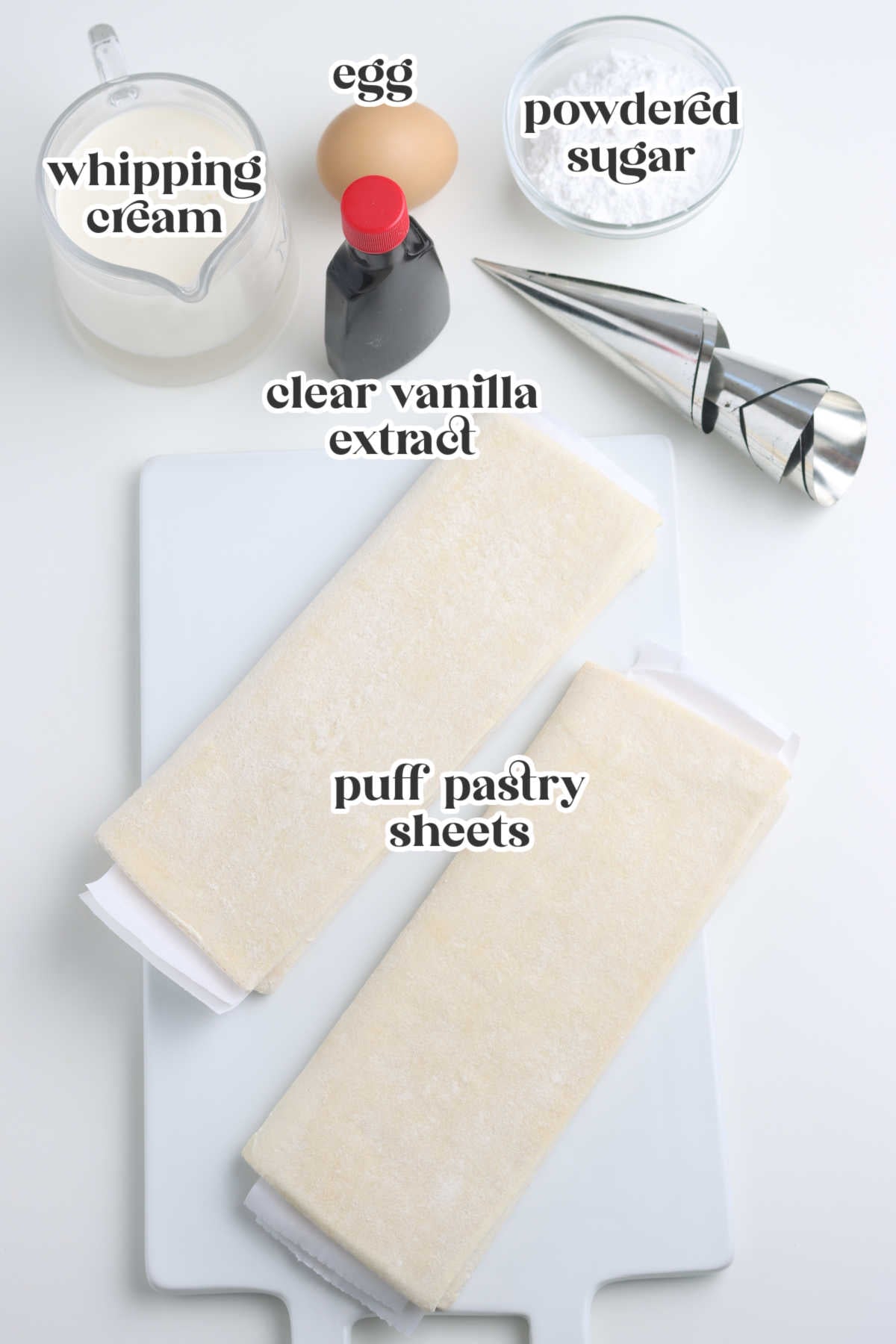 Ingredients for puff pastry cream horns including puff pastry sheets, whipping cream, egg, powdered sugar, and clear vanilla extract displayed on a white cutting board with cream horn molds.