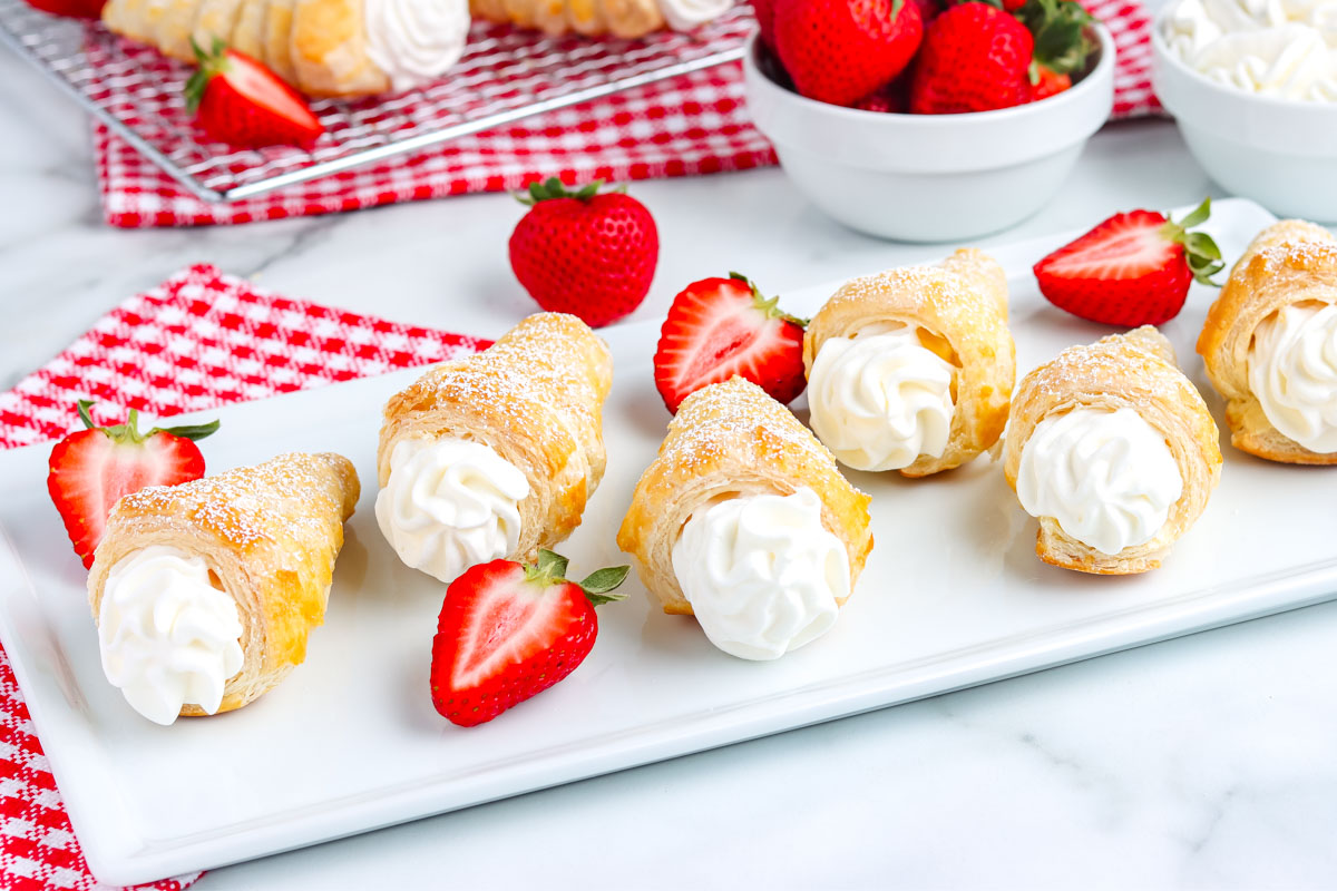 Finished puff pastry cream horns filled with vanilla whipped cream and garnished with fresh strawberries on a white platter with a red checkered napkin.