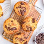Chocolate chunk banana muffins cooling on a wire rack with chopped chocolate and ripe bananas in the background.