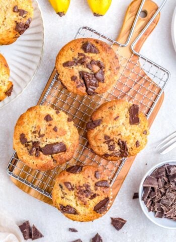 Chocolate chunk banana muffins cooling on a wire rack with chopped chocolate and ripe bananas in the background.