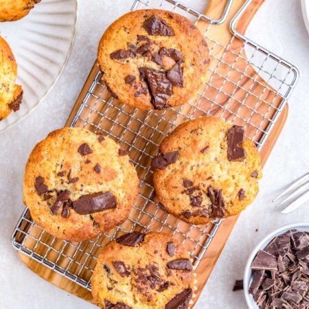 Chocolate chunk banana muffins cooling on a wire rack with chopped chocolate and ripe bananas in the background.