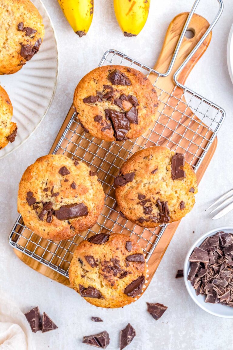 Chocolate chunk banana muffins cooling on a wire rack with chopped chocolate and ripe bananas in the background.