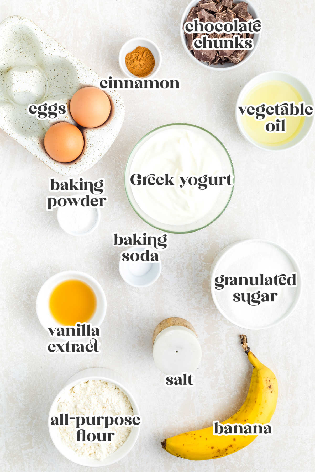 Ingredients for chocolate chunk banana muffins including mashed banana, flour, eggs, Greek yogurt, chocolate chunks, sugar, and cinnamon.