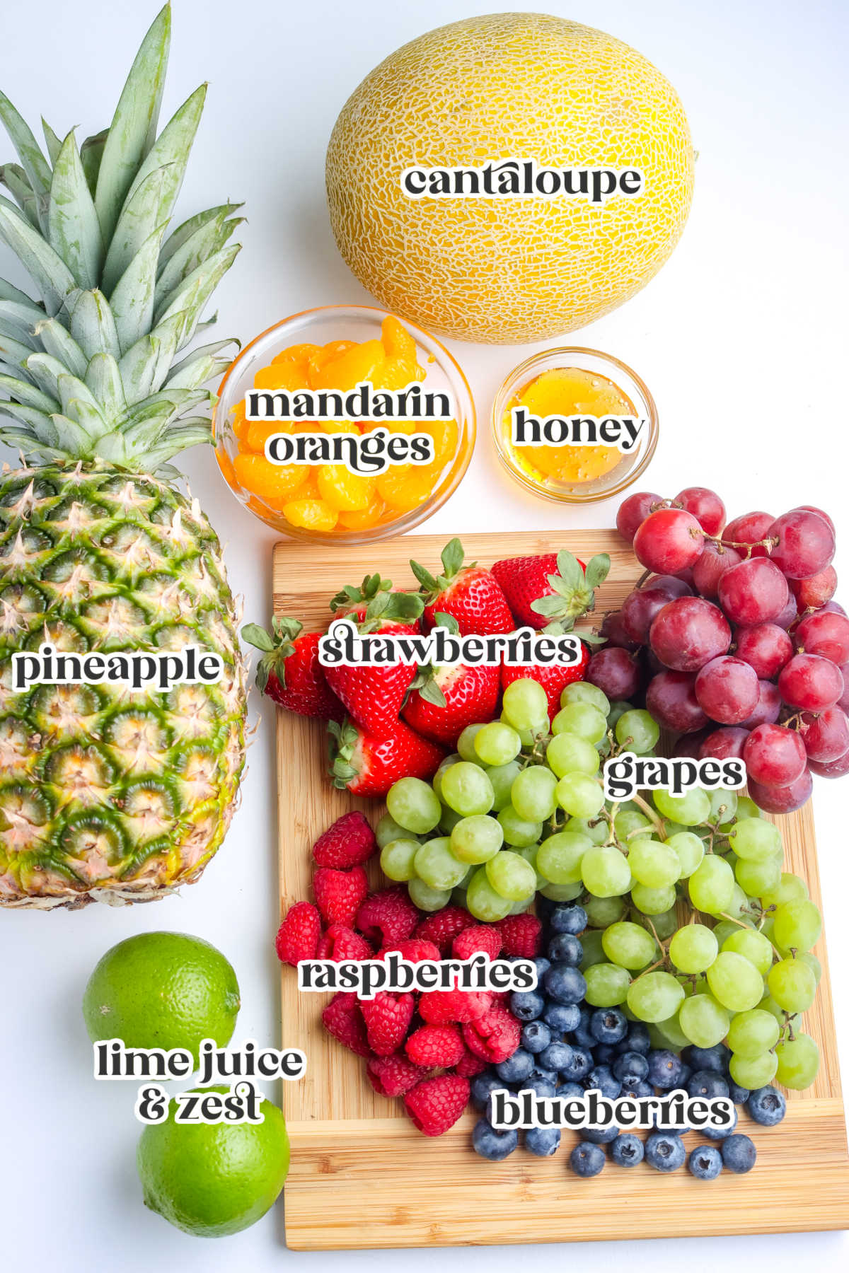 Fresh fruit salad ingredients arranged on a white surface including cantaloupe, pineapple, strawberries, grapes, blueberries, raspberries, mandarin oranges, lime, and honey.