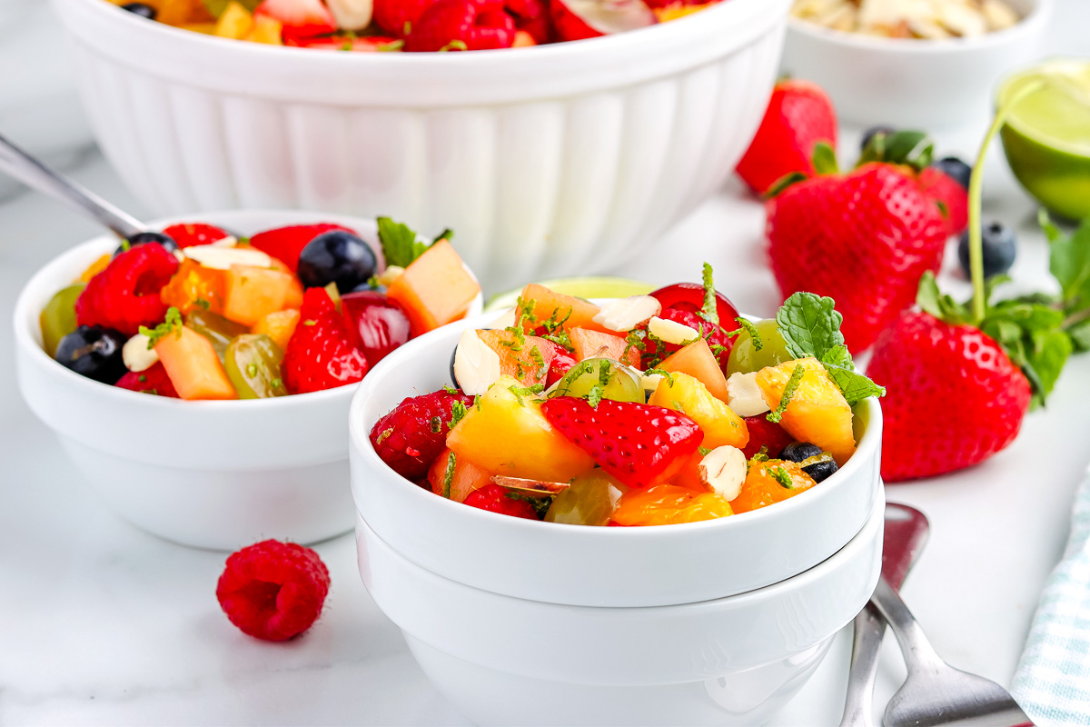 Individual bowls of fruit salad with lime dressing topped with sliced almonds and fresh mint on a bright tabletop.
