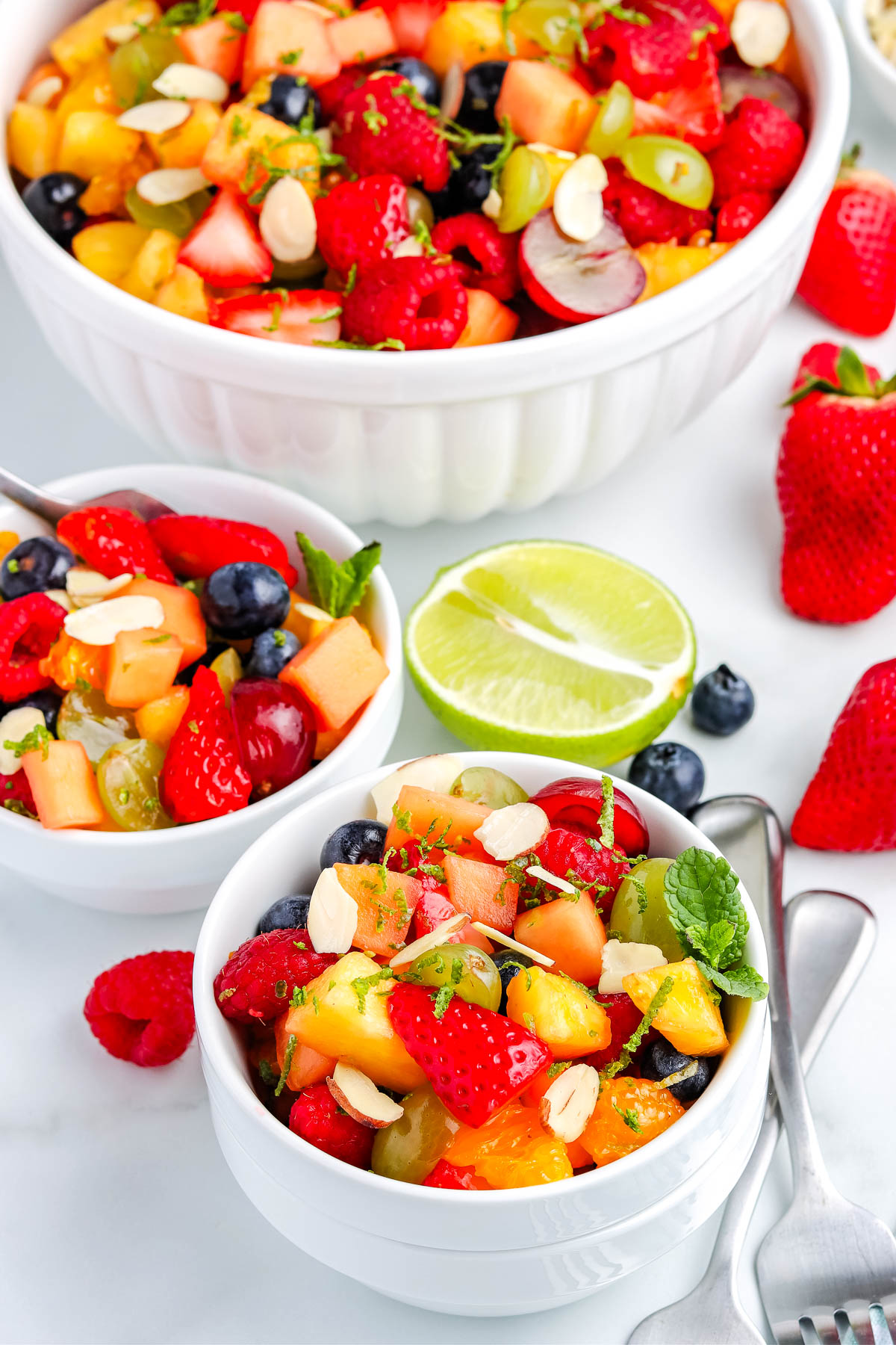 Close up of fruit salad in small white bowls with strawberries, grapes, pineapple, and lime garnish on the side.