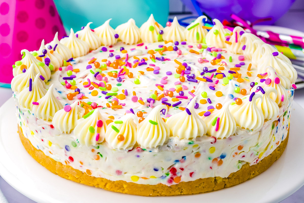 Whole no-bake funfetti cheesecake decorated with piped vanilla icing and colorful sprinkles on a white cake stand.