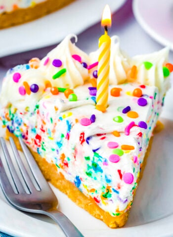 Slice of no-bake funfetti cheesecake with rainbow sprinkles and a birthday candle on a white dessert plate.
