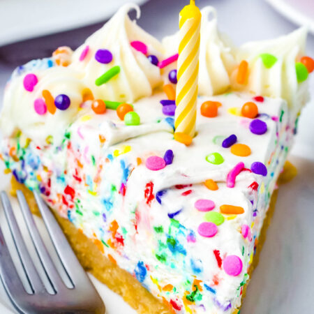 Slice of no-bake funfetti cheesecake with rainbow sprinkles and a birthday candle on a white dessert plate.
