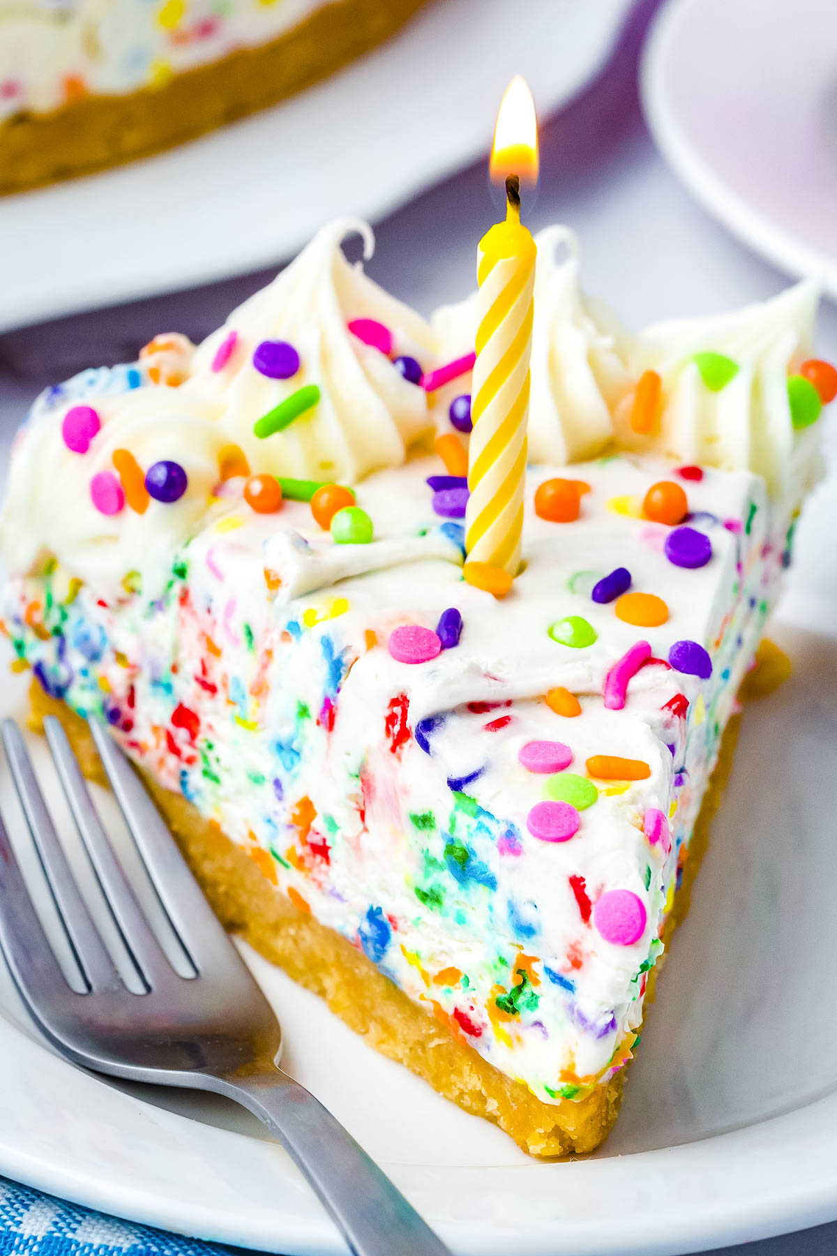 Slice of no-bake funfetti cheesecake with rainbow sprinkles and a birthday candle on a white dessert plate.