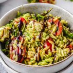 A grey ceramic bowl filled with creamy green summer orzo salad featuring halved cherry tomatoes, mozzarella pearls, baby arugula, and a dark balsamic glaze.