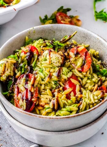 A grey ceramic bowl filled with creamy green summer orzo salad featuring halved cherry tomatoes, mozzarella pearls, baby arugula, and a dark balsamic glaze.