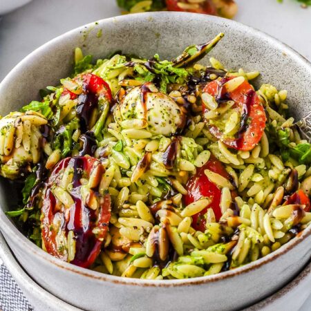 A grey ceramic bowl filled with creamy green summer orzo salad featuring halved cherry tomatoes, mozzarella pearls, baby arugula, and a dark balsamic glaze.