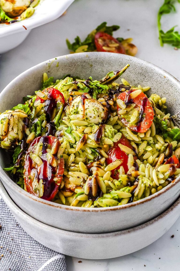 A grey ceramic bowl filled with creamy green summer orzo salad featuring halved cherry tomatoes, mozzarella pearls, baby arugula, and a dark balsamic glaze.