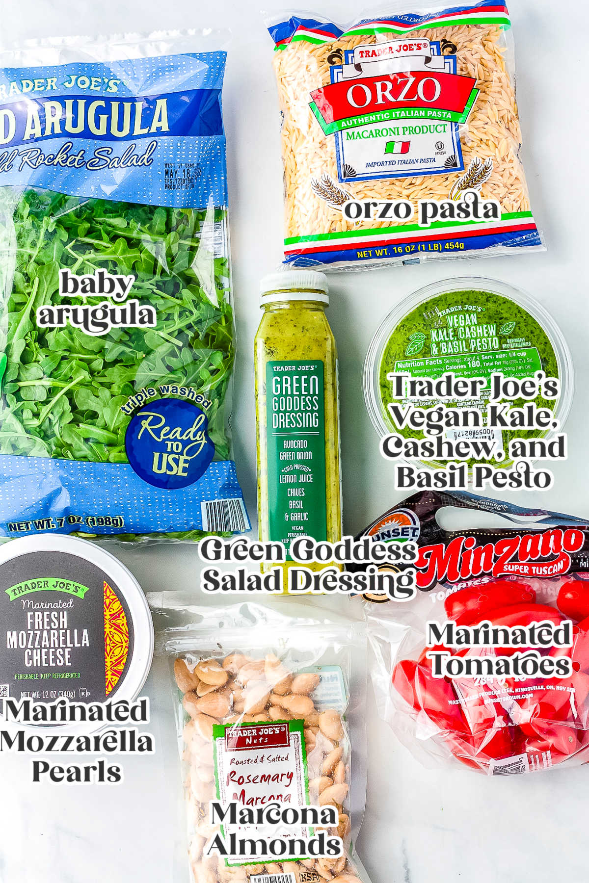 A flat lay showing labeled Trader Joe's ingredients for summer orzo salad, including baby arugula, orzo pasta, Green Goddess salad dressing, vegan pesto, marinated mozzarella pearls, marinated tomatoes, and Marcona almonds.