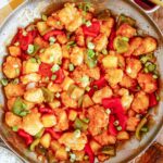An overhead view of a large metal skillet filled with sweet and sour chicken mixed with pineapple and bell peppers. Freshly sliced green onions rest on top of the glossy sauce.