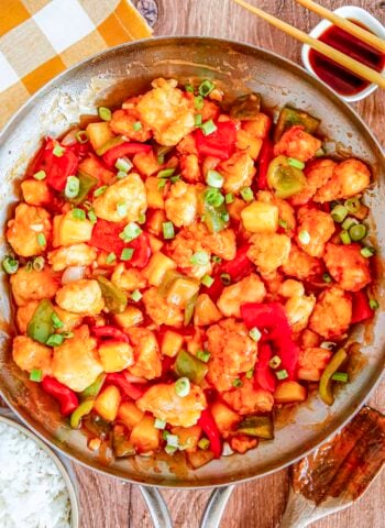 An overhead view of a large metal skillet filled with sweet and sour chicken mixed with pineapple and bell peppers. Freshly sliced green onions rest on top of the glossy sauce.