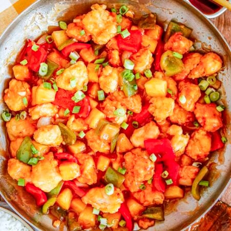 An overhead view of a large metal skillet filled with sweet and sour chicken mixed with pineapple and bell peppers. Freshly sliced green onions rest on top of the glossy sauce.