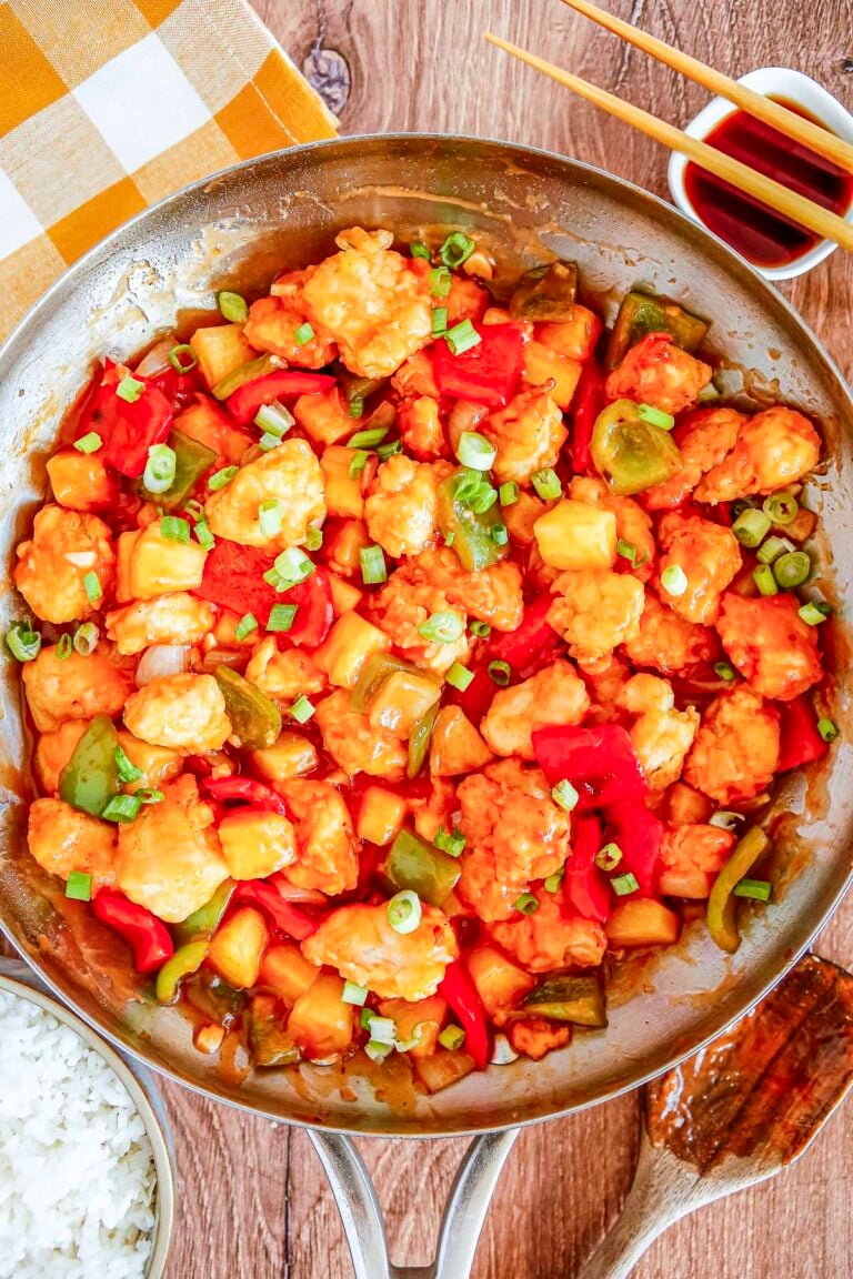 An overhead view of a large metal skillet filled with sweet and sour chicken mixed with pineapple and bell peppers. Freshly sliced green onions rest on top of the glossy sauce.
