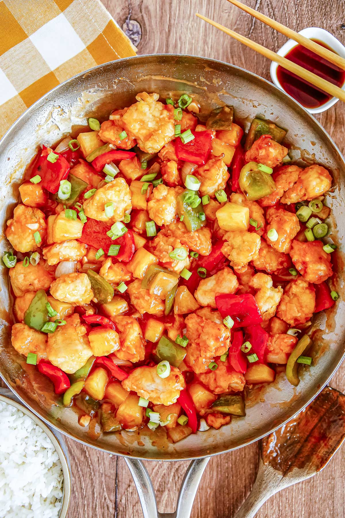 An overhead view of a large metal skillet filled with sweet and sour chicken mixed with pineapple and bell peppers. Freshly sliced green onions rest on top of the glossy sauce.