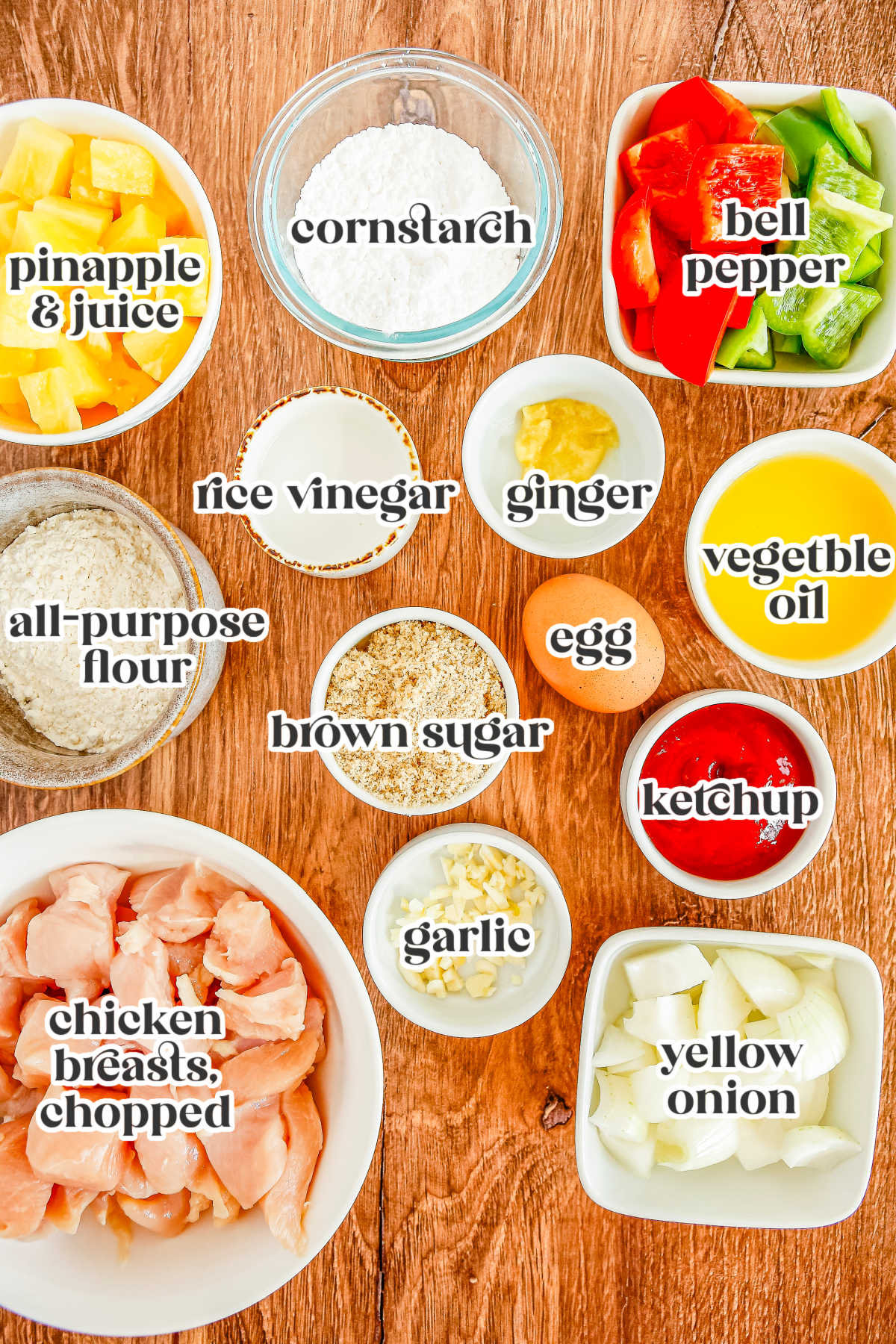 A flat lay of labeled ingredients to make sweet and sour chicken showing pineapple chunks, cornstarch, bell peppers, rice vinegar, ginger, vegetable oil, flour, brown sugar, an egg, ketchup, raw chicken breast, garlic, and a yellow onion.